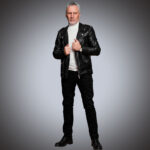 Full-body view of a man wearing a premium black leather biker jacket with silver hardware, styled with a white turtleneck and black trousers, suitable for private label fashion catalogs.