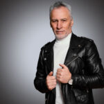 Side profile of a man wearing a high-quality black leather biker jacket with silver metallic hardware, featuring reinforced shoulder epaulettes and a professional slim-fit design.