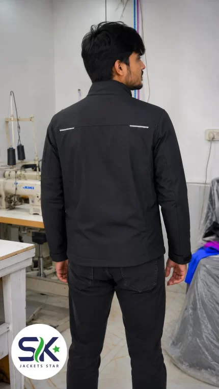 Rear view of model wearing black windproof workwear softshell jacket with reflective back accents in Sialkot apparel manufacturing plant.
