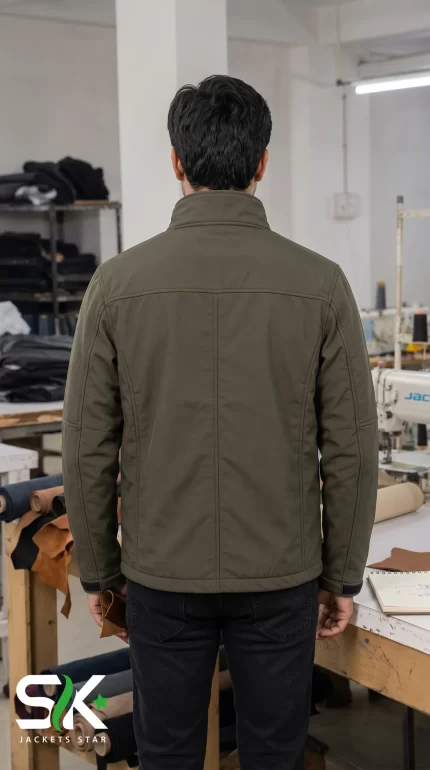 Back view of a male model wearing an olive green softshell tactical jacket in a garment manufacturing facility.