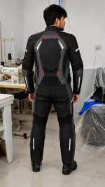 Rear view of a model wearing a black, grey, and red Cordura biker suit with speed hump in a factory setting.